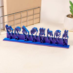 May include: A purple and blue 3D printed sign spelling out the word "THANK" in American Sign Language.