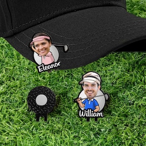 May include: Black golf hat with a personalized ball marker featuring a cartoon image of a person with the name "Eleanor". Also, a black golf ball marker and a personalized marker with the name "William". The markers are on a green grass background.