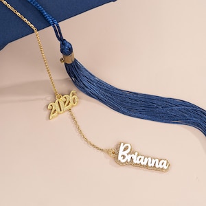 May include: A navy blue graduation tassel with a gold chain and charms. The charms include the year "2026" and the name "Brianna" in a white and gold design. The tassel is hanging from a navy blue graduation cap.