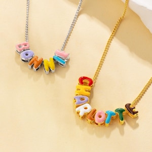 May include: Two necklaces featuring name pendants. One has a silver chain with the name "BONNIE" in pastel-coloured letters. The other has a gold chain with the name "CHARLOTTE" in vibrant, multi-coloured letters.