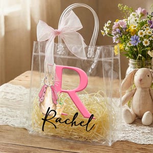 May include: Clear tote bag with a pink bow and handles, featuring a pink letter "R" and the name "Rachel" in script. A bunny illustration is on the front. The bag is filled with yellow straw.