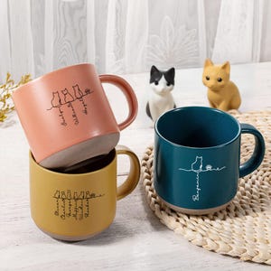 Puede incluir: Tres tazas de cerámica en melocotón, amarillo y verde azulado, cada una con un diseño de silueta de gato y nombres. Las tazas tienen un diseño de dos tonos con una base marrón claro y un asa curva. Dos figuras de gato están en el fondo.