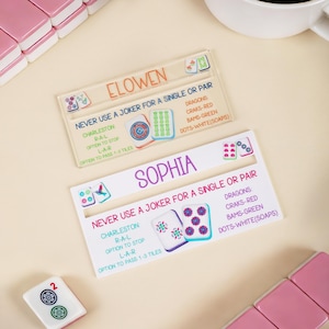 May include: Two personalized Mahjong tile holders with names, text, and game instructions. The holders are white and clear with colorful illustrations of Mahjong tiles. The name "ELOWEN" is in orange, and "SOPHIA" is in purple.
