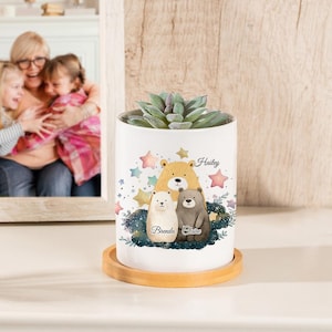 May include: White ceramic planter with a wood base. The planter features a watercolor illustration of three bears with the names Hailey, Brenda, and Claire. The planter is filled with a green succulent plant.