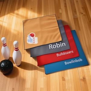 May include: Four square microfiber bowling towels in tan, gray, red, and blue. The tan towel features a bowling ball and pins graphic. The gray towel says Robin, the red towel says Bulldozers, and the blue towel says Bowlinbob in white text.