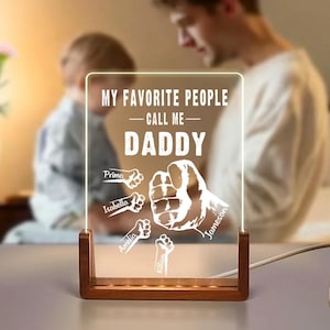 May include: A personalized LED night light with a wooden base. The clear acrylic panel displays the text "MY FAVORITE PEOPLE CALL ME DADDY" along with fist and hand illustrations and names. The light is powered by a white cord.