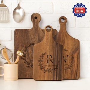 Könnte beinhalten: Drei Holzschneidebretter mit Griffen. Die Bretter sind mit floralen Designs und personalisiertem Text graviert. Das mittlere Brett trägt den Namen "ROWAN" und den Text "Love You Forever". Hergestellt in den USA.