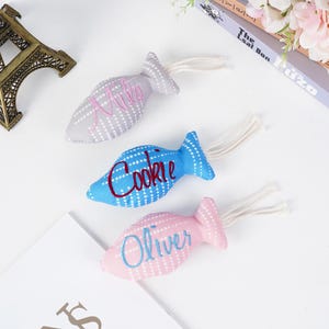 May include: Three fish-shaped cat toys in gray, blue, and pink, each with a different name embroidered on them. The toys have white polka dots and a white string tail. The names are "Milo", "Cookie", and "Oliver".