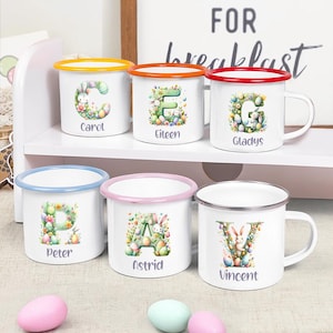 May include: Six white enamel mugs with colorful rims and handles are displayed on a white shelf. Each mug features a letter and name, decorated with Easter-themed illustrations of bunnies, eggs, and flowers. The mugs have yellow, orange, red, blue, pink, and silver rims.