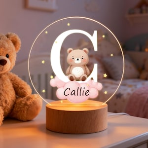 May include: A personalized night light featuring the letter "G" with a teddy bear illustration. The name "Callie" is written below the bear on a pink cloud. The light sits on a wooden base and is surrounded by star-shaped accents.