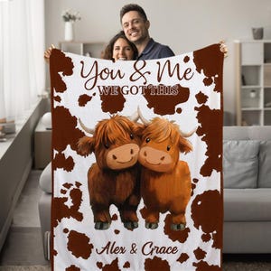May include: White and brown fleece blanket with a cowhide pattern. Two cartoon Highland cows are cuddling. The text "You & Me We Got This" is printed on the blanket. The names "Alex & Grace" are printed at the bottom of the blanket.