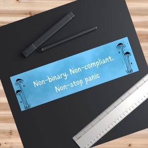 May include: A rectangular light blue sticker featuring black line art of mushrooms and stars on the left and right sides. The text reads, "Non-binary, Non-compliant, Non-stop panic." A black stylus and a ruler are positioned near the sticker.