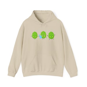 Puede incluir: Una sudadera con capucha beige con un gráfico de tres lúpulos verdes de dibujos animados con caras sonrientes. Los lúpulos sostienen una regadera azul.