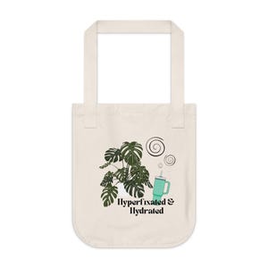 Puede incluir: Bolso tote de color crema con una ilustración de una planta de monstera verde, una taza turquesa y el texto "Hyperfixated & Hydrated". El bolso tiene dos asas y un fondo redondeado. El diseño incluye espirales.