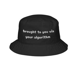 以下が含まれることがあります： 白い文字で「brought to you via your algorithm」と書かれた黒いバケットハット。つばが広く、丸みを帯びたクラウンが特徴です。柔らかくしなやかな素材で作られています。ユニセックスアクセサリー。