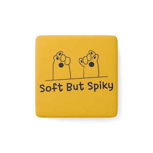 Puede incluir: Un imán cuadrado amarillo con el texto "Soft But Spiky" en negro. Encima del texto hay dos manos de dibujos animados con palmas negras. Las manos están dibujadas en negro.