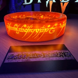 May include: Glowing orange ring with engraved text, resting above a business card. The ring's inscription is illuminated, and the card displays the text "Z COOKED DESIGNS" in a graffiti style.