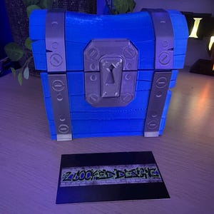 May include: A blue treasure chest with silver accents, including hinges and a keyhole. The chest is made of layered material and sits on a wooden surface. A business card with the text "ZLOOKED DESIGNS" is in front of the chest.