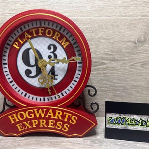 Puede incluir: Un reloj rojo y dorado con las palabras "PLATFORM 9 3/4" y "HOGWARTS EXPRESS". El reloj tiene manecillas doradas y una base negra con volutas. A la derecha hay una tarjeta de visita negra con texto estilo grafiti.
