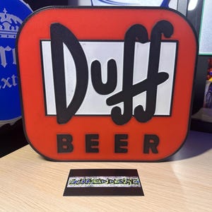 Pode incluir: Uma placa vermelha e branca da Duff Beer com letras pretas. A placa é retangular com cantos arredondados. A palavra "Duff" está em letras grandes e em negrito, e "BEER" está escrito abaixo. Um cartão de visita com o texto "2LOOKED DESIGNS" está em frente à placa.