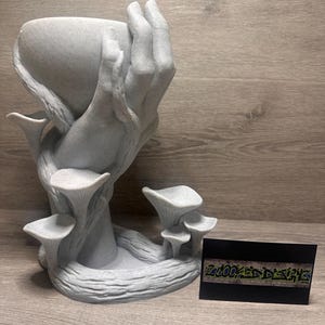 Op de afbeelding: Een grijze, 3D-geprinte plantenbak in de vorm van een hand die een kom vasthoudt, met paddenstoelachtige structuren en een getextureerde basis. Een visitekaartje met graffiti-achtige tekst staat naast de plantenbak.