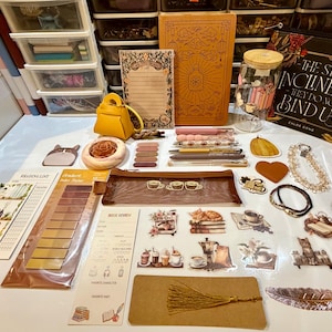 Puede incluir: Una colección de artículos de papelería y accesorios, que incluye un bolso amarillo, cuadernos, pegatinas, bolígrafos y un libro titulado "The Stars Incline Us They Do Not Bind Us". Los artículos están dispuestos sobre una superficie blanca.