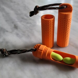 May include: Orange earplug containers with a textured surface and a black and grey cord. One container is open, revealing two green earplugs. The other two containers are closed. The containers are cylindrical and have a dotted pattern.