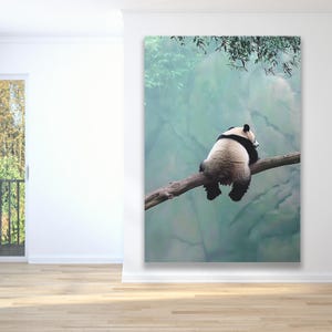 May include: A black and white panda bear sits on a branch with its back to the camera. The panda is looking to the right and has its head resting on the branch. The background is a blurry green and blue.