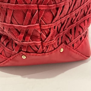 May include: Close-up of a red leather handbag with a woven design. The bag features a series of interwoven leather strips, creating a textured pattern. Gold-colored studs are visible on the base of the bag.