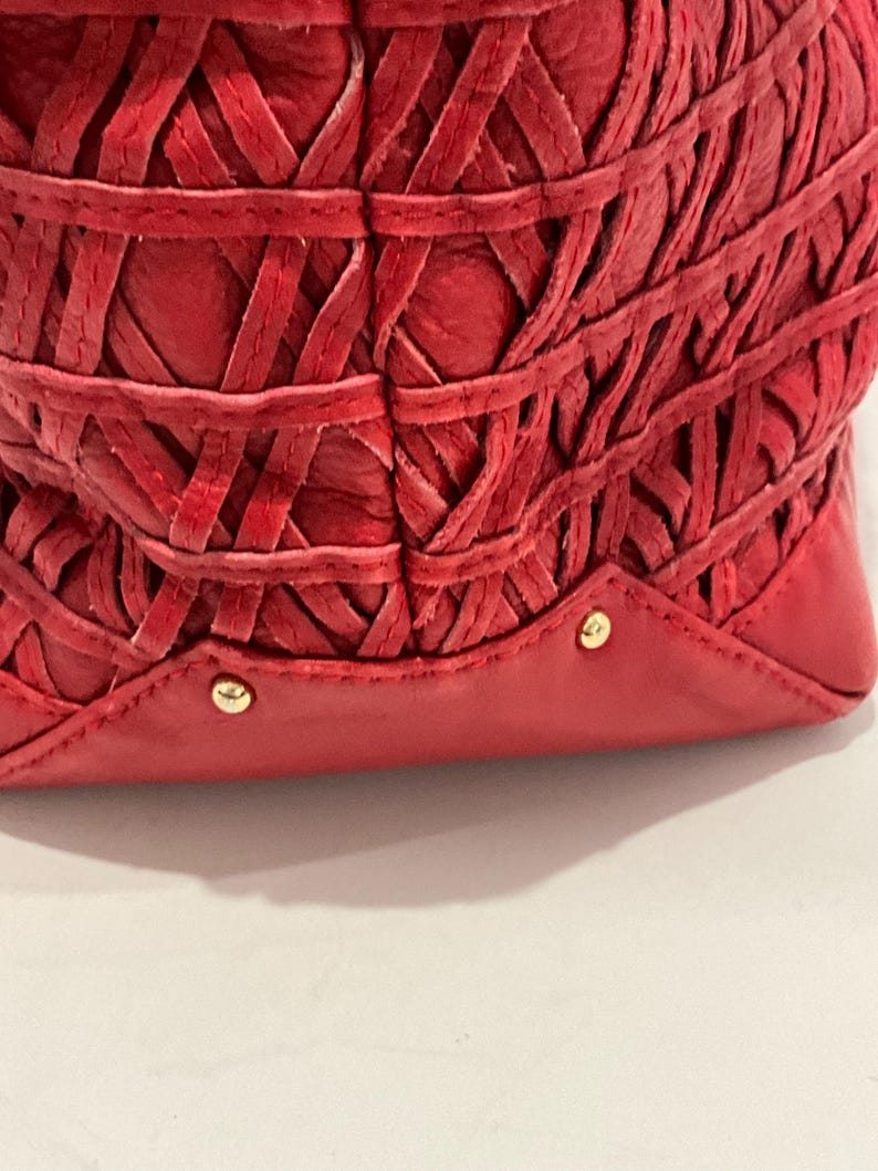 May include: Close-up of a red leather handbag with a woven design. The bag features a series of interwoven leather strips, creating a textured pattern. Gold-colored studs are visible on the base of the bag.