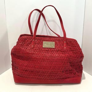 May include: A red leather tote bag with woven detailing and two top handles. The bag features a rectangular shape with a flat base and a gold-colored rectangular plaque on the front. The bag is designed for carrying items.