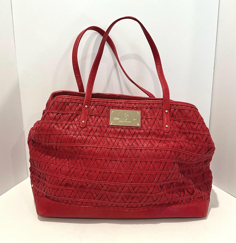 May include: A red leather tote bag with woven detailing and two top handles. The bag features a rectangular shape with a flat base and a gold-colored rectangular plaque on the front. The bag is designed for carrying items.