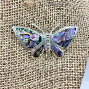 Puede incluir: Un broche de mariposa de plata con alas de nácar de abulón iridiscente. Las alas tienen una gama de colores púrpura, verde y azul.