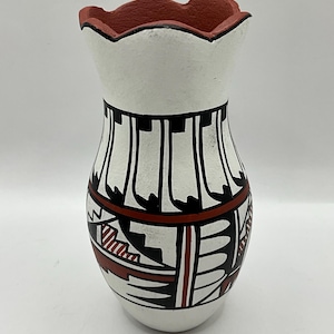 Puede incluir: Un jarrón de cerámica blanca con un borde festoneado y un cuerpo acampanado. El jarrón presenta patrones geométricos negros y rojos y líneas verticales negras. El borde está pintado de rojo.