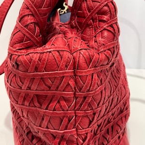 May include: A red leather handbag with a woven design. The bag features a structured base with gold-colored studs and a soft, gathered top. The strap is a thin, matching red leather.
