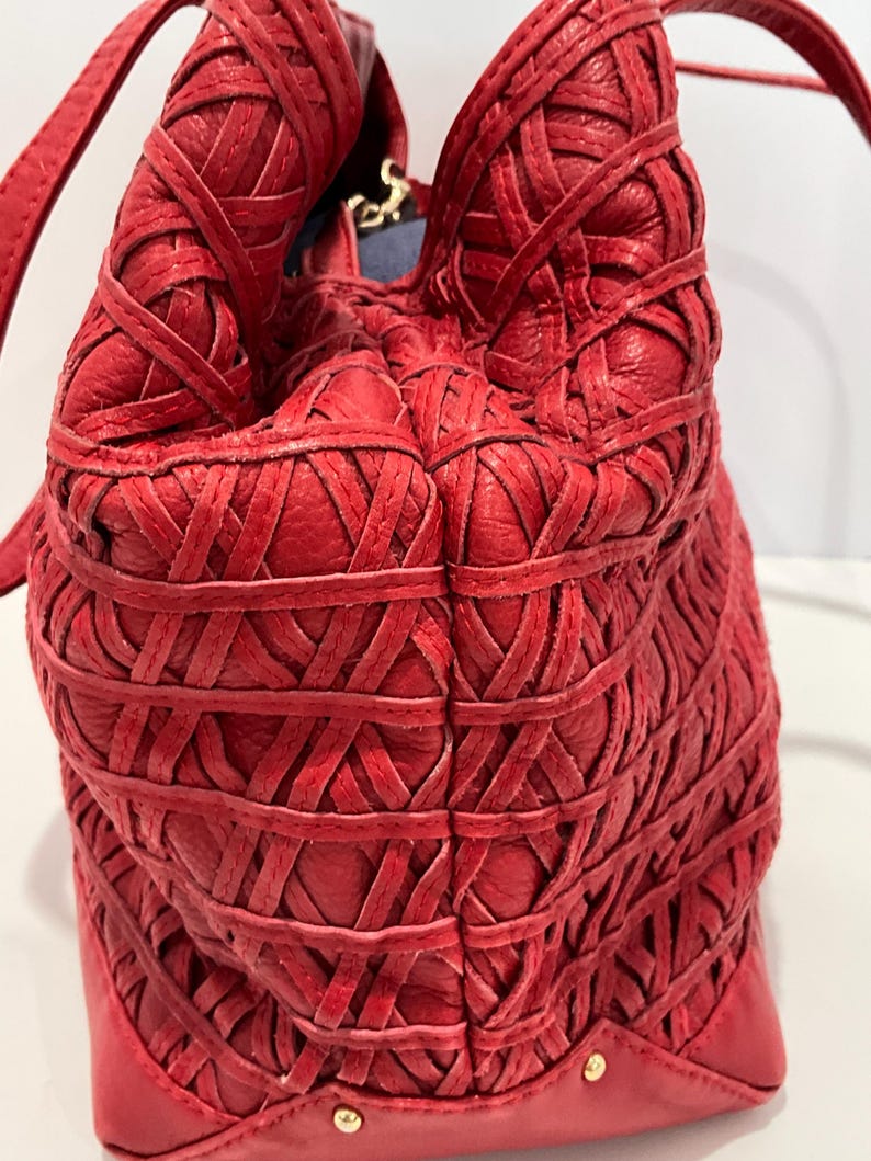 May include: A red leather handbag with a woven design. The bag features a structured base with gold-colored studs and a soft, gathered top. The strap is a thin, matching red leather.