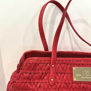 May include: A red tote bag with woven detailing and two top handles. The bag features a gold-colored rectangular plaque with a logo. The bag is made of a textured material.