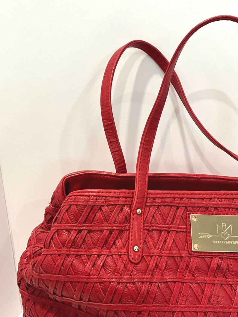 May include: A red tote bag with woven detailing and two top handles. The bag features a gold-colored rectangular plaque with a logo. The bag is made of a textured material.