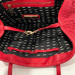 May include: Open view of a red leather tote bag. The bag features a woven exterior design and a black interior with a white dash pattern. It has a zippered pocket and a gold-colored clasp.