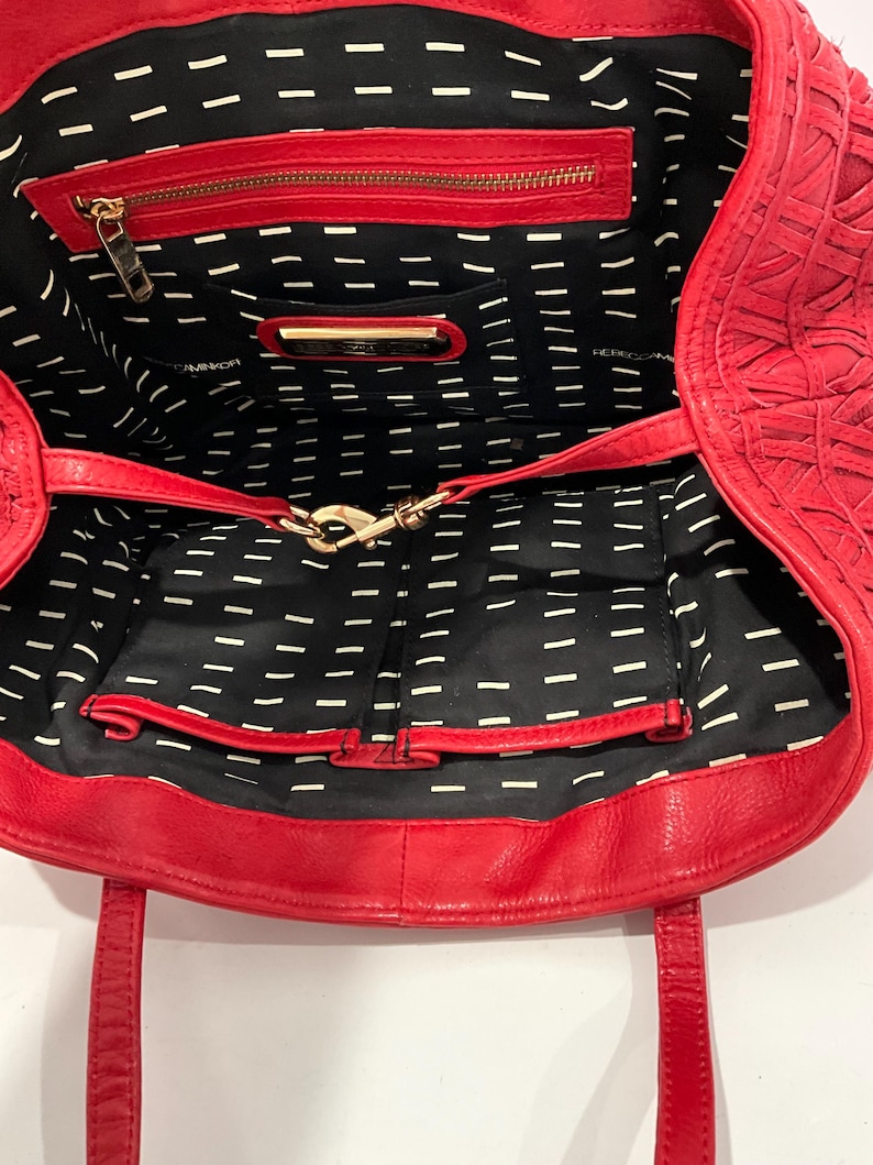 May include: Open view of a red leather tote bag. The bag features a woven exterior design and a black interior with a white dash pattern. It has a zippered pocket and a gold-colored clasp.