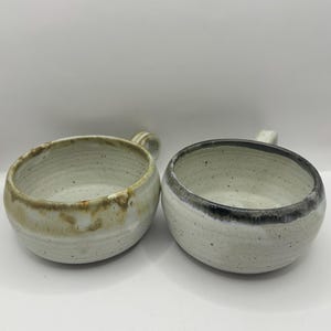 May include: Two handmade ceramic bowls with handles. One bowl has a light beige and brown rim, the other has a dark grey rim. Both bowls are round with a speckled, off-white interior.