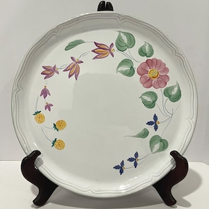 Op de afbeelding: Witte keramische serveerschaal met een geschulpte rand, versierd met handgeschilderde bloemen- en fruitontwerpen in roze, paars, geel en groen. De schaal staat op een donkere houten standaard.