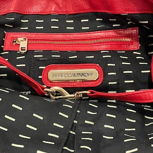May include: Red leather handbag with a black interior featuring a zipper pocket and a gold-tone metal logo that reads "REBECCAMINKOFF". The bag has a red strap and a black lining with white dashes.