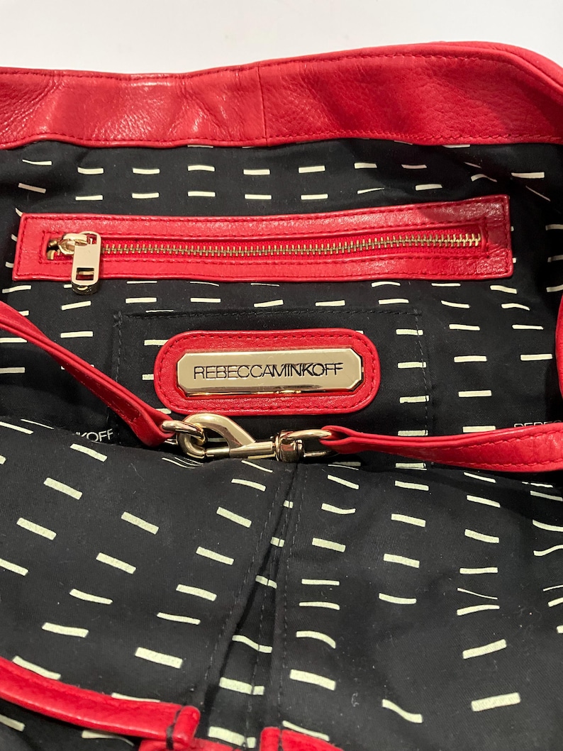May include: Red leather handbag with a black interior featuring a zipper pocket and a gold-tone metal logo that reads "REBECCAMINKOFF". The bag has a red strap and a black lining with white dashes.