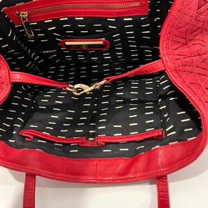 May include: Red leather tote bag with an open interior. The bag features a zippered pocket and a gold-tone clasp. The interior lining is black with a white dash pattern. The bag has two handles.