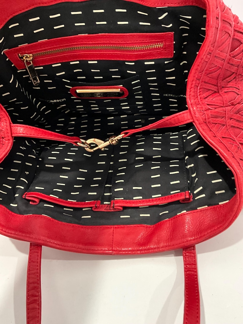 May include: Red leather tote bag with an open interior. The bag features a zippered pocket and a gold-tone clasp. The interior lining is black with a white dash pattern. The bag has two handles.