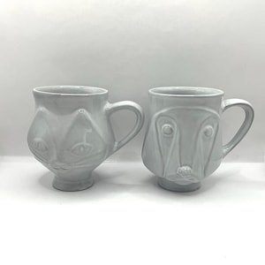 Può includere: Due tazze in ceramica bianca. Una tazza presenta un disegno a forma di gatto, l'altra a forma di cane. Entrambe le tazze hanno un manico e una base arrotondata. Le tazze sono su uno sfondo bianco.