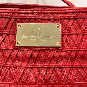 May include: Close-up of a red leather handbag with a woven design. A gold-colored rectangular plaque is centered on the bag, featuring an arrow, heart, and text. The bag has a textured surface and visible stitching.