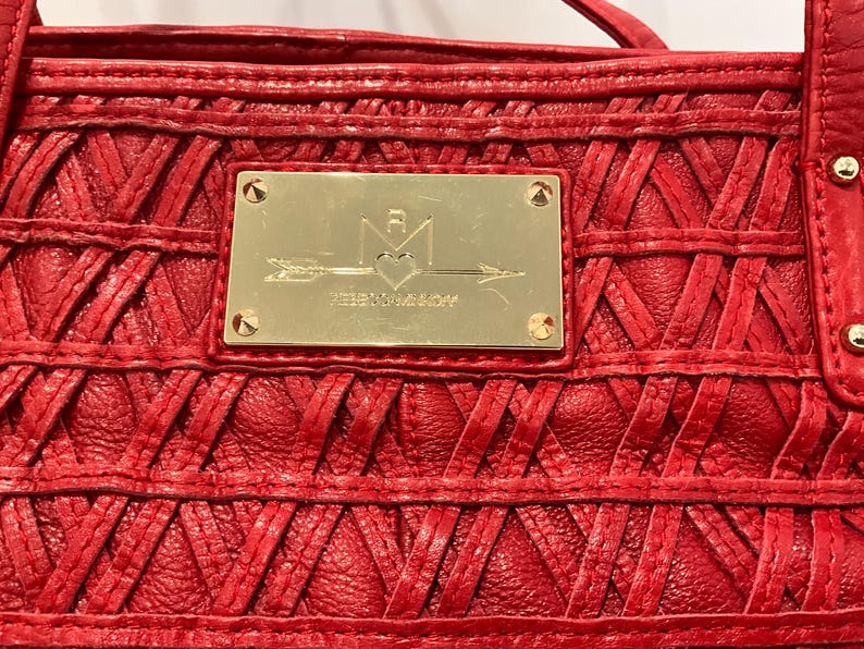 May include: Close-up of a red leather handbag with a woven design. A gold-colored rectangular plaque is centered on the bag, featuring an arrow, heart, and text. The bag has a textured surface and visible stitching.