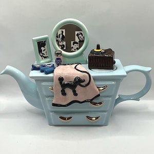 以下が含まれることがあります： 水色のセラミックティーポット。ドレッサーのデザインで、黒いプードルが描かれたピンクのタオル、額入りの写真、画像付きの小さな鏡が特徴です。 ティーポットにはハンドルと注ぎ口が付いています。